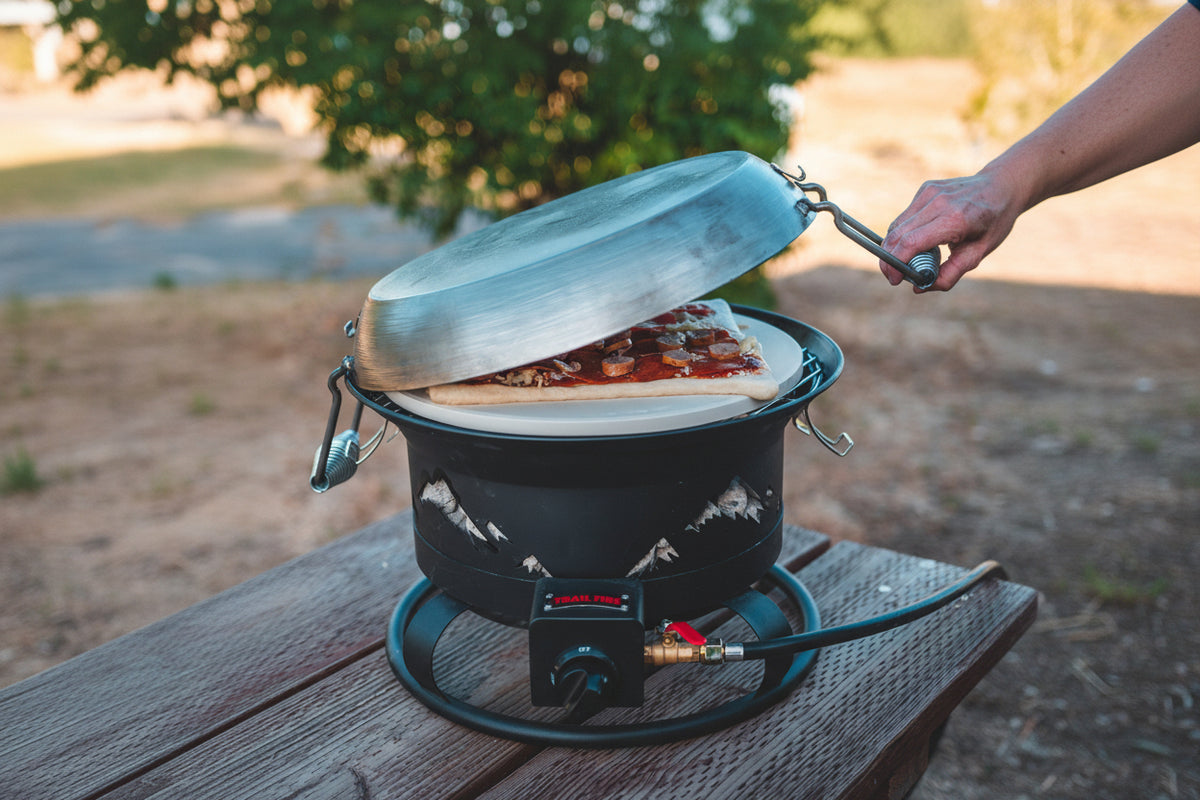 Portable pizza oven with a pizza being cooked outdoors on a wooden surface.